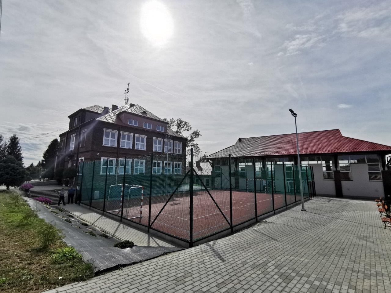 Za ogrodzonym boisko z nawierzchnią tartanową, budynek szkoły po lewej, a po prawej hala sportowa.