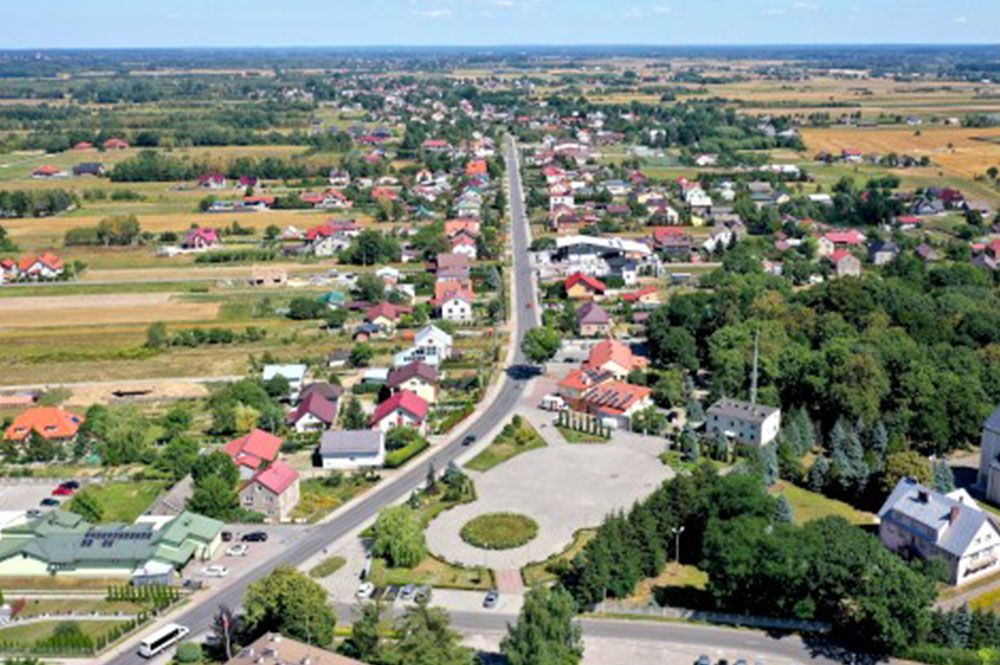 Panorama wsi z lotu ptaka. W centrum plac oraz budynki, po lewej i prawej stronie pola i drzewa.